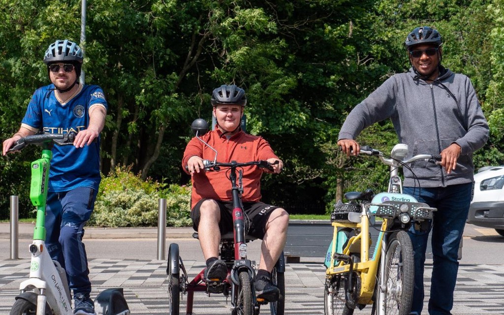 The image is of three men using micromobility equipment, one e-scooter, one cycle and one e-bike, outside in a park. The sun is shining.
