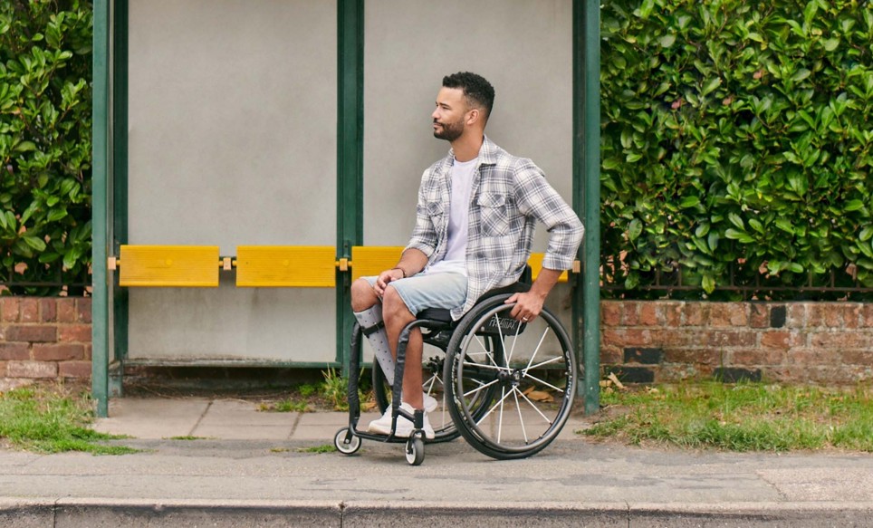Jordan is a wheelchair user at is parked at a bus stop. Behind Jordan is a bus shelter and big green hedge.