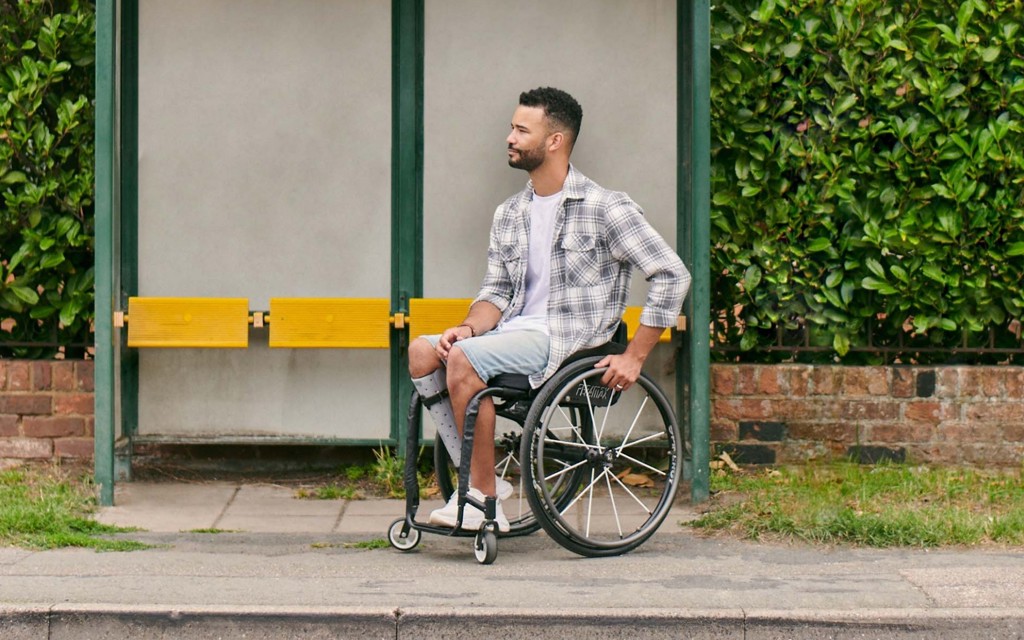Jordan is a wheelchair user at is parked at a bus stop. Behind Jordan is a bus shelter and big green hedge.