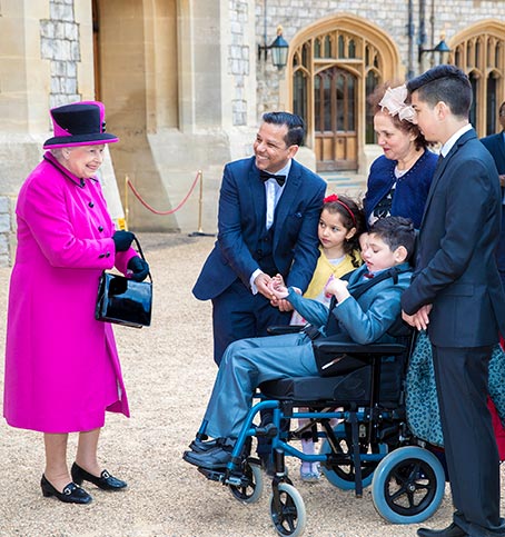 Her majesty the queen is in a pink coat and hat, she is smiling and standing next to a family. The family are smiling at her majesty.