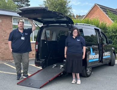 A min-bus with writing on the side of the vehicle reads Worcester Wheels. The mini-bus is facing away with the boot open and the ramp down. On the left-hand side of the open boot is a man with short brown and blonde hair wearing a Worchester Wheels branded navy polo, green trousers and black trainers. On the right-hand side of the open boot is a women with long brown hair wearing a Worchester Wheels branded navy polo, black skirt and pink trainers.