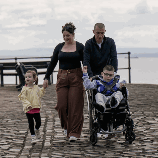 Gino is being wheeled along a cobbled path near the sea by his dad. Walking next to them is his mum and sister. They are talking to one another.