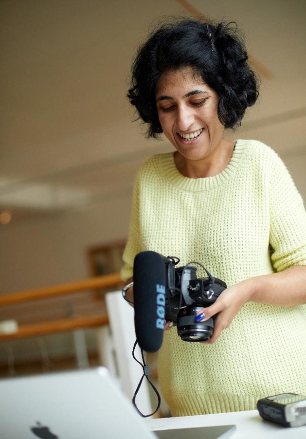 Mathy is smiling whilst looking down at the screen on her photography camera. Next to her is a digital camera and her laptop on a white table. She is wearing a yellow knitted jumper and has short black hair in a bob.