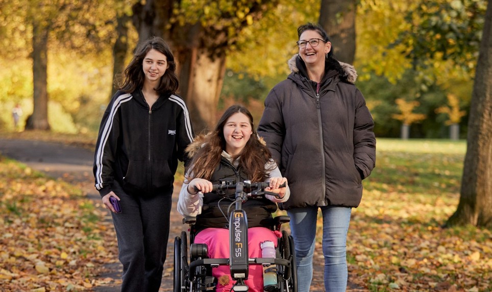 Becky, Holly and Sarah are in their local park, the leaves on the many trees around them are yellow and orange. Becky is wheeling in her wheelchair in the middle, whilst her sister Holly is to her left and her mum Sarah is to the right. 
