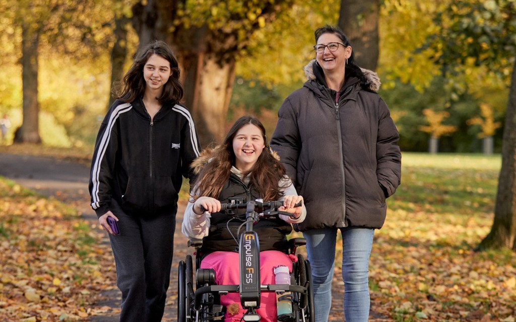 Becky, Holly and Sarah are in their local park, the leaves on the many trees around them are yellow and orange. Becky is wheeling in her wheelchair in the middle, whilst her sister Holly is to her left and her mum Sarah is to the right. 