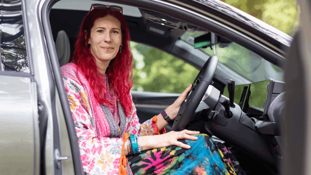 Charlie has pink hair and is wearing multi coloured clothing. They are sitting in their silver vehicle facing the camera smiling.