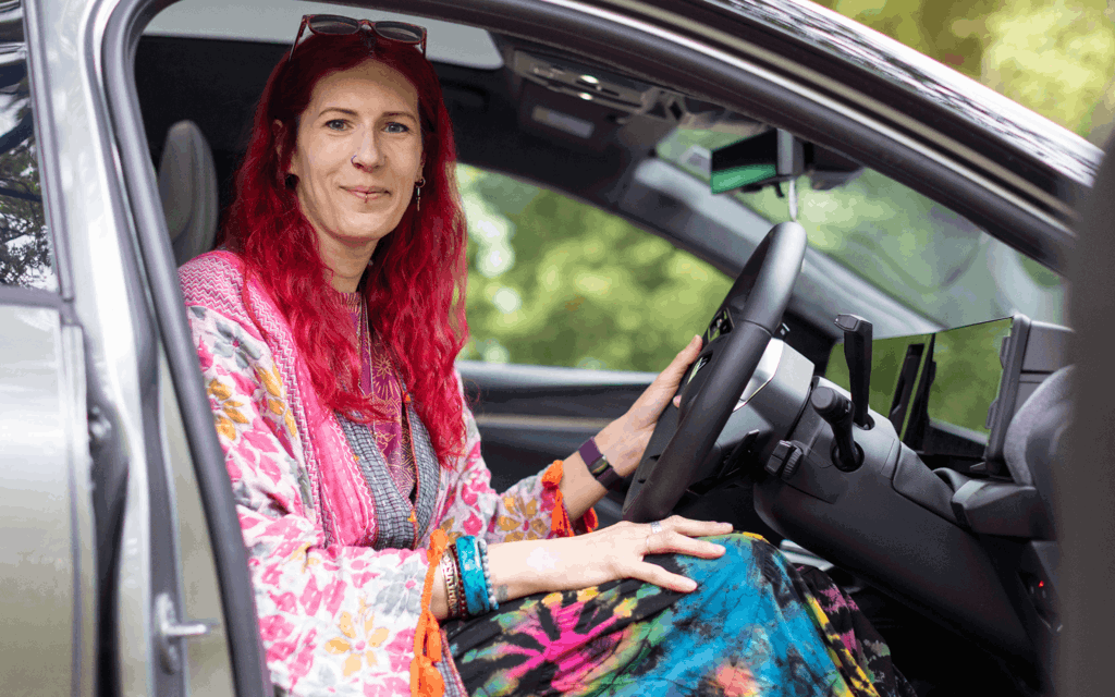 Charlie has pink hair and is wearing multi coloured clothing. They are sitting in their silver vehicle facing the camera smiling.