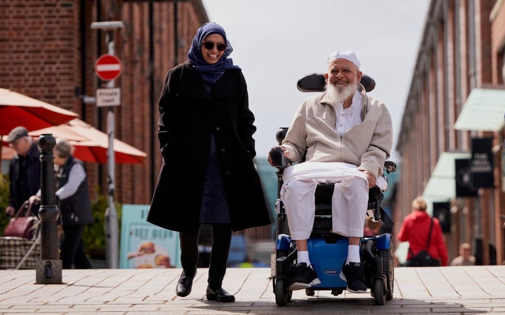 The image has red bricked buildings in the background with shops and big umbrellas from outside restaurant seating. Moosa's wife is on the left-hand side walking and talking with Moosa who is smiling on the right-hand side in his electric wheelchair. 