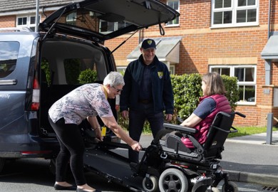The image shows a gentleman in a navy blue baseball cap, navy blue jacket, black trousers and black trainers standing at the back of a vehicle with the ramp down smiling. An older lady smiling with grey hair, patterned pink top, black trousers and black ballerina pumps is to the right of him attaching a wheelchair to the ramp. In the centre of the image is a women seated in the wheelchair with brown hair, wearing a bright pink gilet.
