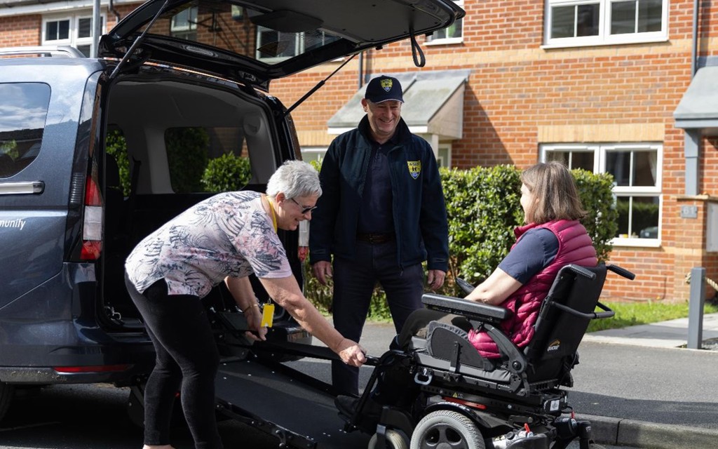 The image shows a gentleman in a navy blue baseball cap, navy blue jacket, black trousers and black trainers standing at the back of a vehicle with the ramp down smiling. An older lady smiling with grey hair, patterned pink top, black trousers and black ballerina pumps is to the right of him attaching a wheelchair to the ramp. In the centre of the image is a women seated in the wheelchair with brown hair, wearing a bright pink gilet.