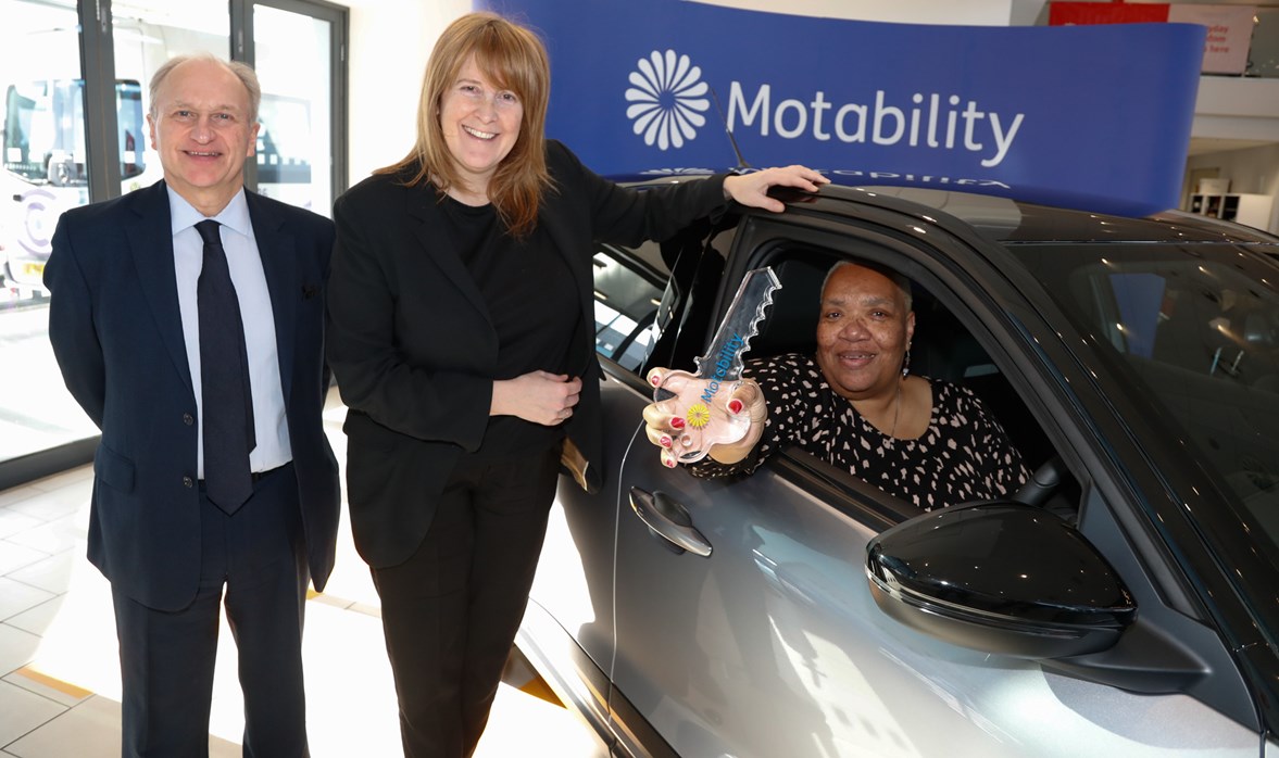 A smiling man and woman stand next to another woman sitting in a shiny new car holding a big key