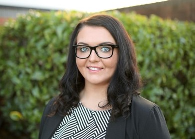 Emma Partlow ncat CEO is smiling in a black blazer and white and black stripped top. She has long brown curly hair and black rimmed glasses.