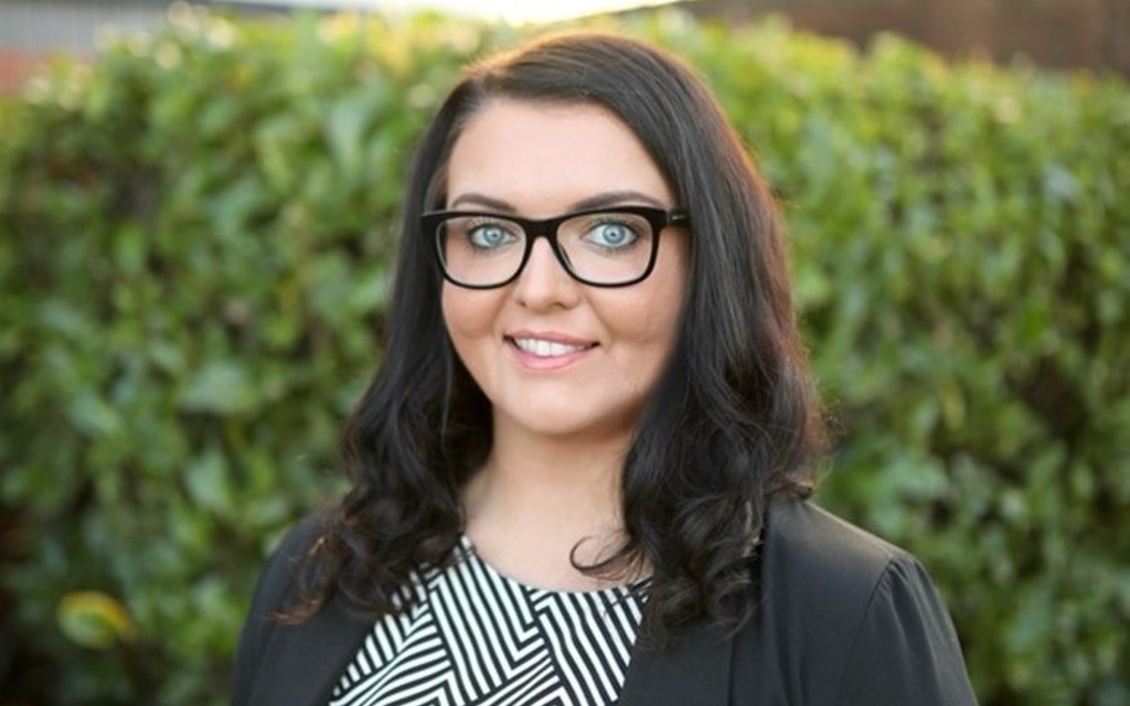 Emma Partlow ncat CEO is smiling in a black blazer and white and black stripped top. She has long brown curly hair and black rimmed glasses.