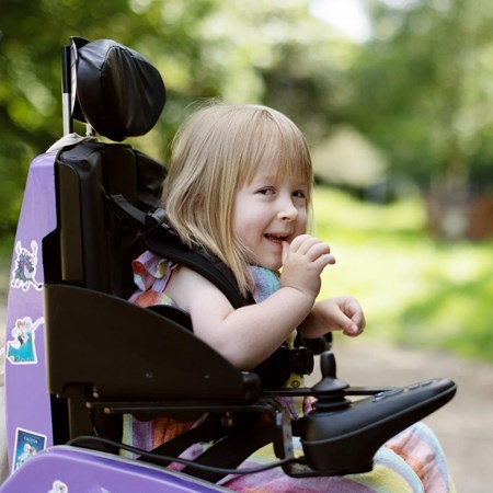 The image is a close up of Poppy in her lilac powered wheelchair. She is laughing. Behind her are lots of green trees. 