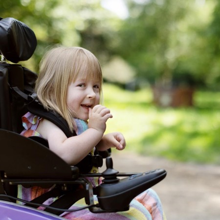 The image is a close up of Poppy in her lilac powered wheelchair. She is laughing. Behind her are lots of green trees. 