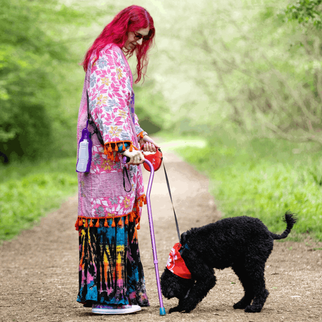Charlie has pink hair and is wearing multi coloured clothing whilst using a pink walking stick. They are holding their dog by the lead on a walk outside. Their dog is black poodle cross and is wearing a red neckerchief with a "in training" sign on their lead.