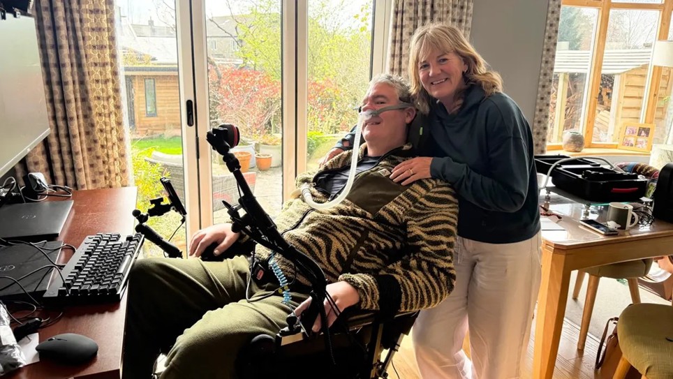 Andrew Herbert is sitting in a powered wheelchair with a women standing behind him.
