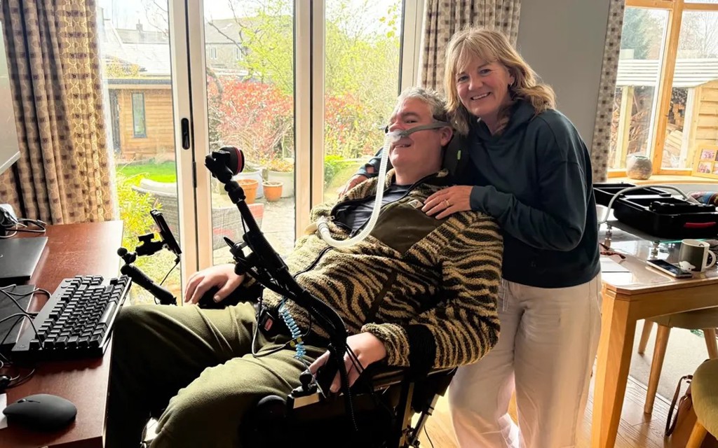 Andrew Herbert is sitting in a powered wheelchair with a women standing behind him.
