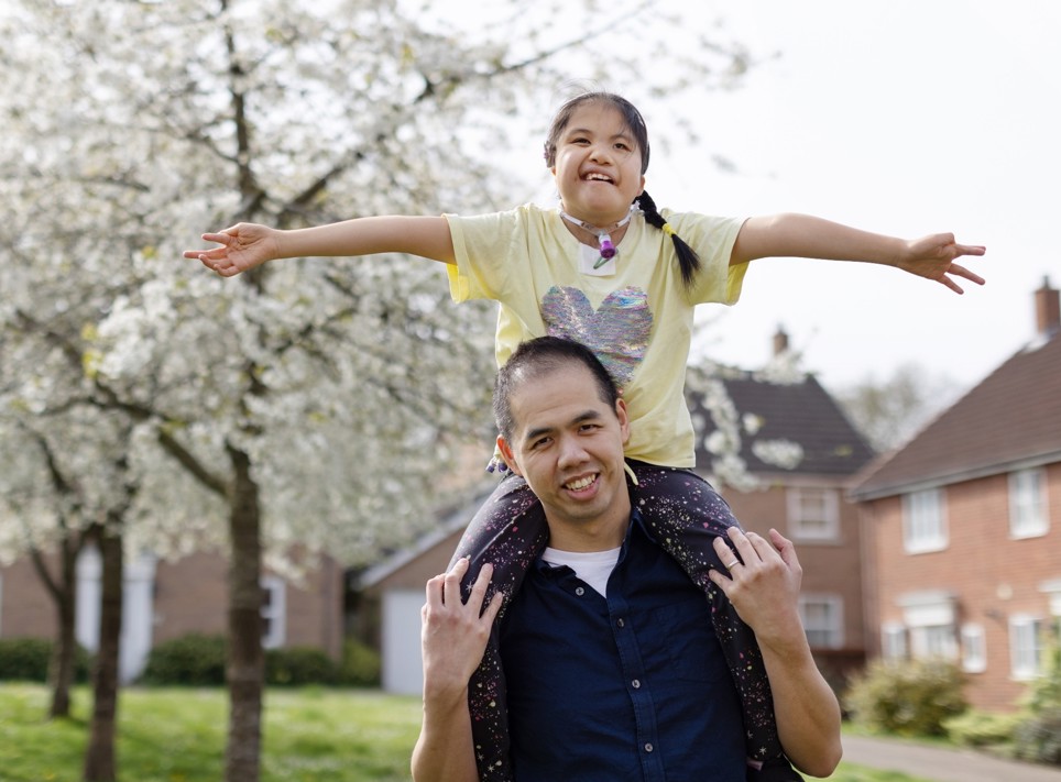 Eva is smiling at the camera in a yellow top, pink headband, she is sitting on her dad's shoulders with her arms stretched wide. Eva's dad is in a navy blue button up short sleeved shirt and smiling.