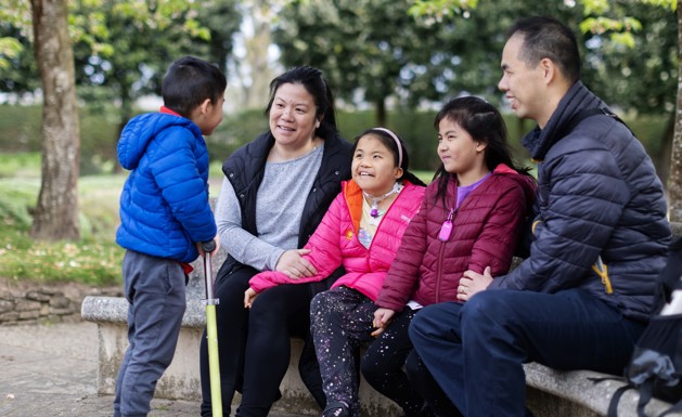 Eva is sitting in the centre of a grey brick wall in a pink puffer coat, pink headband, night sky patterned trousers. Sitting on her right-hand side is her mum and brother. Whereas sitting on her left-hand side is her sister and dad. Everyone is smiling and enjoying being outside.