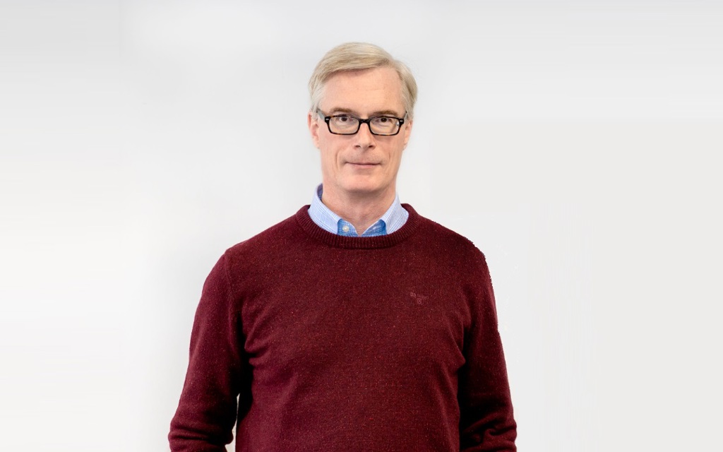 Charles is smiling, he is wearing black rimmed glasses, a red jumper and blue shirt. 