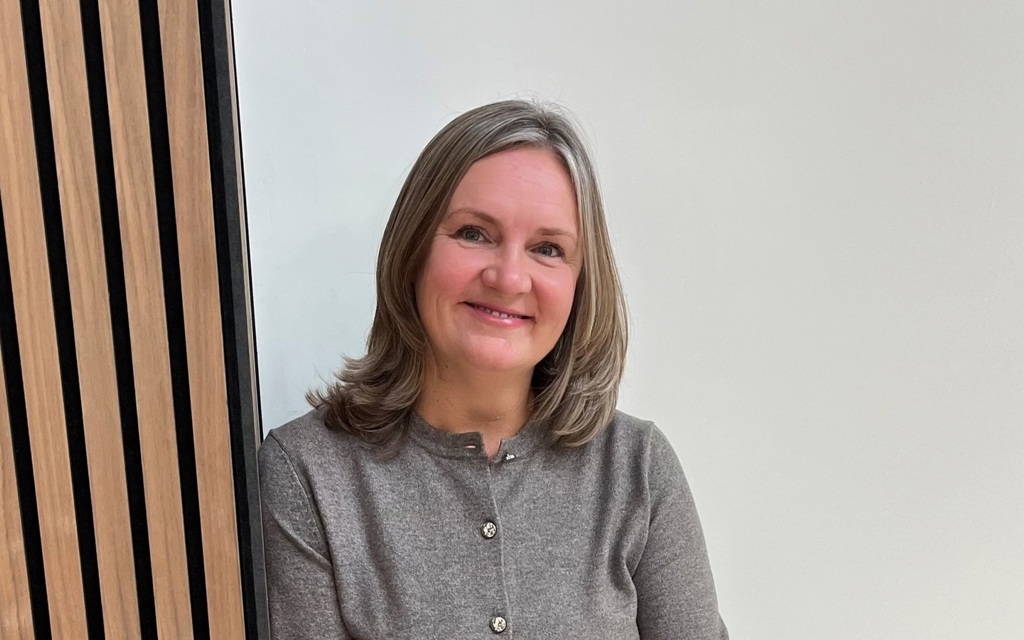 Sam has shoulder length blonde hair, she is smiling at the camera, wearing a grey cardigan.