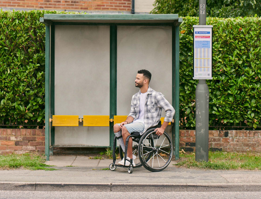 A gentleman is in a wheelchair at a bus stop waiting for a bus to arrive. 