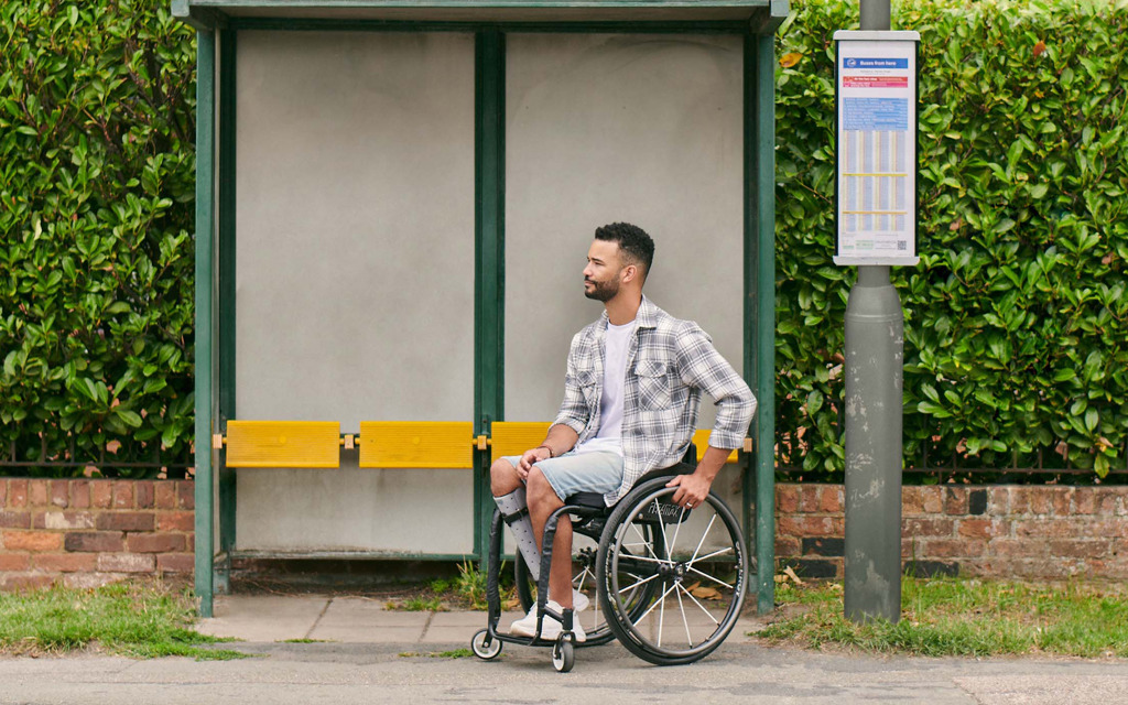 A gentleman is in a wheelchair at a bus stop waiting for a bus to arrive. 